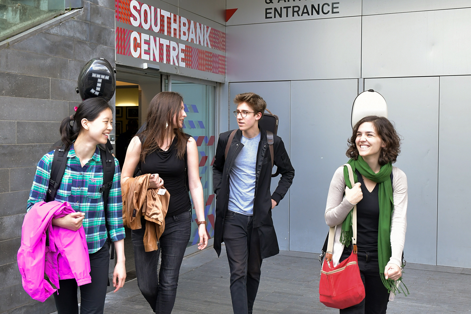 Four students, carrying a bags and music instruments, smiling and chatting, walking outside of the Southbank centre.